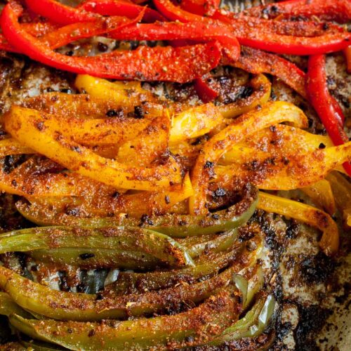 Close-up of roasted brown crispy seasoned red, yellow, green bell pepper strips after baking with tongs taking a couple of them.