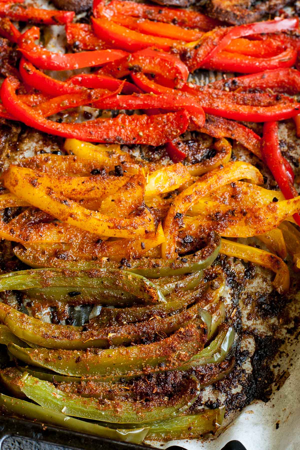 Close-up of roasted brown crispy seasoned red, yellow, green bell pepper strips after baking with tongs taking a couple of them.