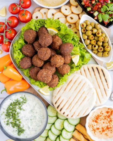 Lot of items in a white board including falafel balls on top of lettuce leaves, charcoaled pita breads, grated cucumber in yoghurt sauce, orange hummus, olives, chopped green red salad, light brown sauce, cherry tomatoes, orange peppers, sliced cucumber, breadsticks and crackers.