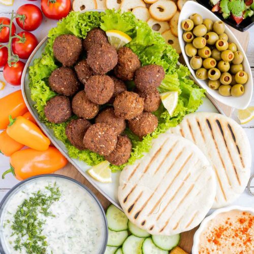 Lot of items in a white board including falafel balls on top of lettuce leaves, charcoaled pita breads, grated cucumber in yoghurt sauce, orange hummus, olives, chopped green red salad, light brown sauce, cherry tomatoes, orange peppers, sliced cucumber, breadsticks and crackers.
