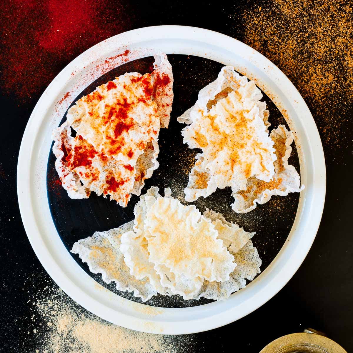 Several puffed rice paper sheets sprinkled with different spices red yellow and brownish on a black plate and those spices are also sprinkled in the black board for contrast