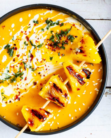 Yellow cream soup topped in a black bowl with charred pineapple wedges, drizzled with a white sauce and sprinkled with chopped green herbs, red flakes and sesame seeds.