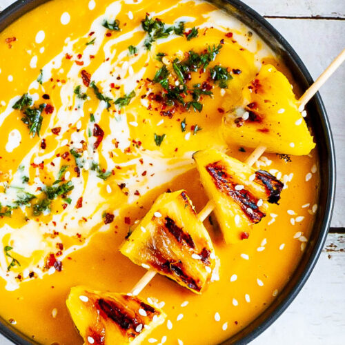 Yellow cream soup topped in a black bowl with charred pineapple wedges, drizzled with a white sauce and sprinkled with chopped green herbs, red flakes and sesame seeds.