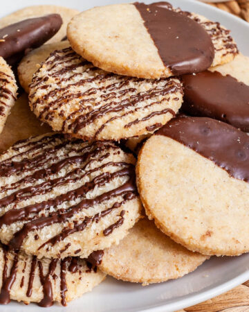 A white plate is full of thin round cookies placed on top of each other. Some cookies are partly covered in chocolate, some cookies are drizzled with chocolate so brown stripes can be seen.