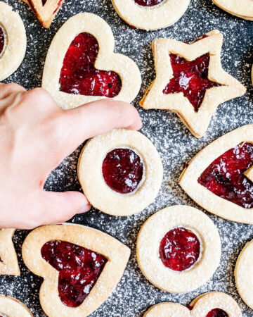 Light brown cookies in shapes of heart, star and circle filled with red jam is scattered around dusted with powdered sugar on a black surface. A hand is taking one cookie from the middle.