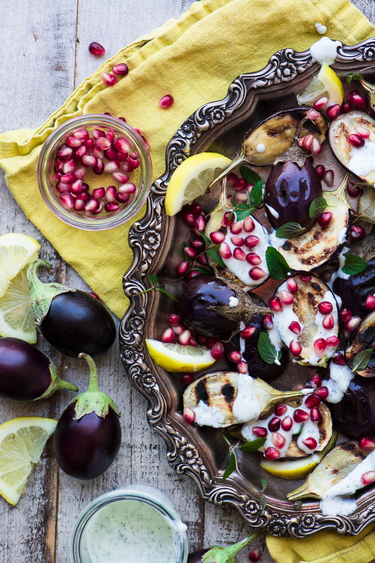 Grilled Baby Eggplant with Minted Yogurt and Pomegranate on a silver tray with napkin and a bowl of pomegranate seeds.
