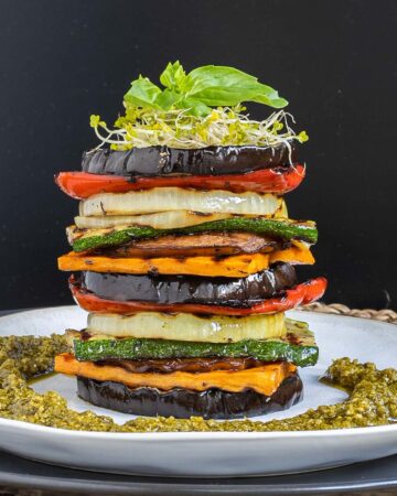 Different veggie slices are stack on top of each other to create a tower of veggies. It has eggplant, zucchini, sweet potato, onion, red bell pepper and mushroom slices. Green pesto is served around it. It is topped with sprouts and basil leaves.
