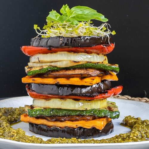 Different veggie slices are stack on top of each other to create a tower of veggies. It has eggplant, zucchini, sweet potato, onion, red bell pepper and mushroom slices. Green pesto is served around it. It is topped with sprouts and basil leaves.