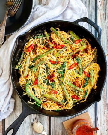 Black cast iron skillet with spaghetti in creamy light brown sauce with asparagus, spinach and red pepper slices. Red harissa sauce in a glass jar and a spoon with tahini paste next to it.