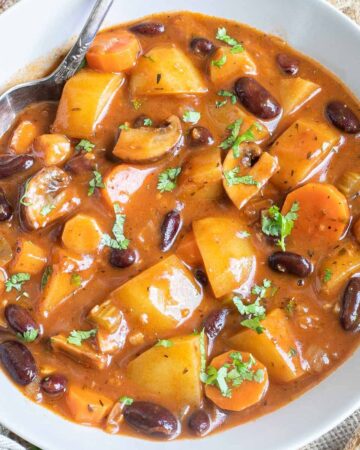 Vegan stew in a white bowl with lots of chopped vegetables like potatoes, carrots, mushroom, celery and red kidney beans. Topped with freshly chopped green herbs.