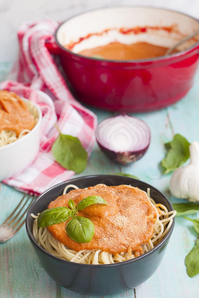 A bowl with vegetable sauce on top of spaghetti sprinkled with vegan parmesan and topped with fresh basil leaves. In the background red Dutch Oven with remaining pasta sauce