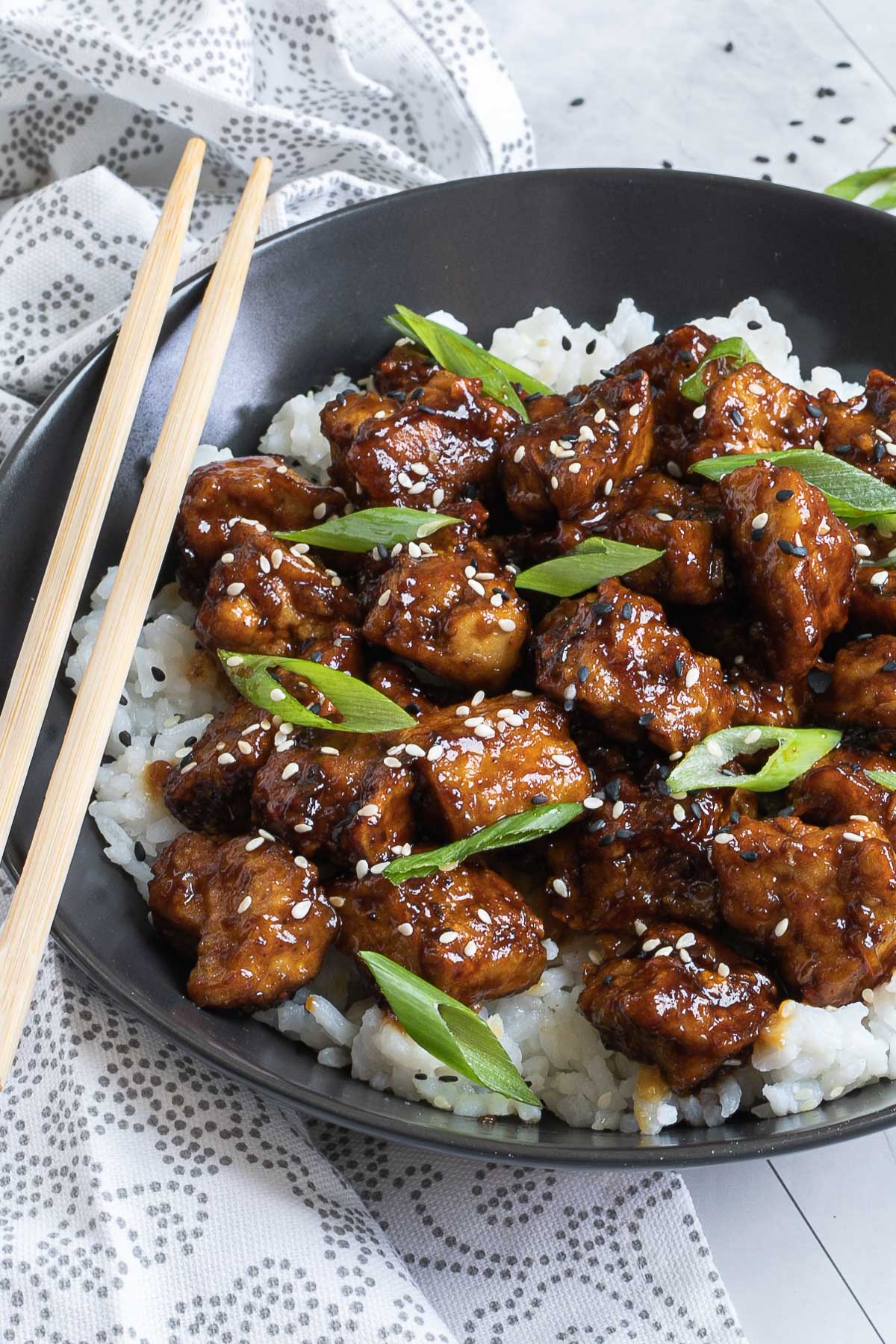Brown sticky tofu pieces served on top of white rice on a black plate. It is sprinkled with black and white sesame seeds and scallion.