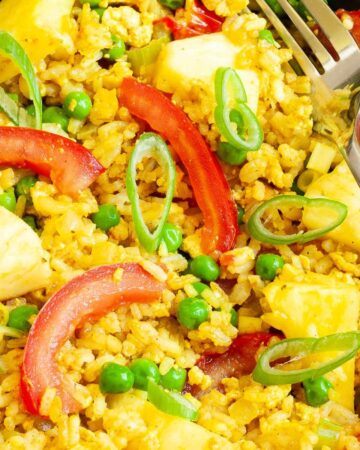 A black bowl with yellow rice, red tomato slices, pineapple chunks, green peas and chopped green onion.