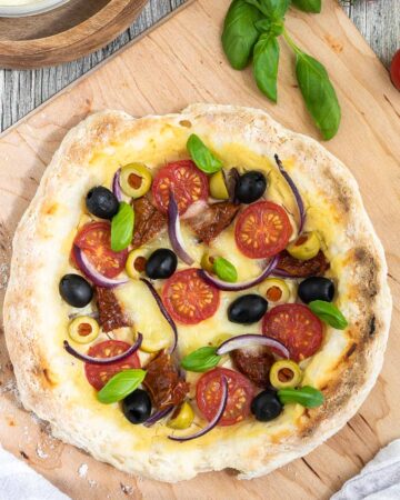 Pizza on a wooden baker's peel topped with light brown hummus, cherry tomatoes, green and black olives, red onion slices, and fresh basil leaves. More hummus and other fresh ingredients are scattered around it.