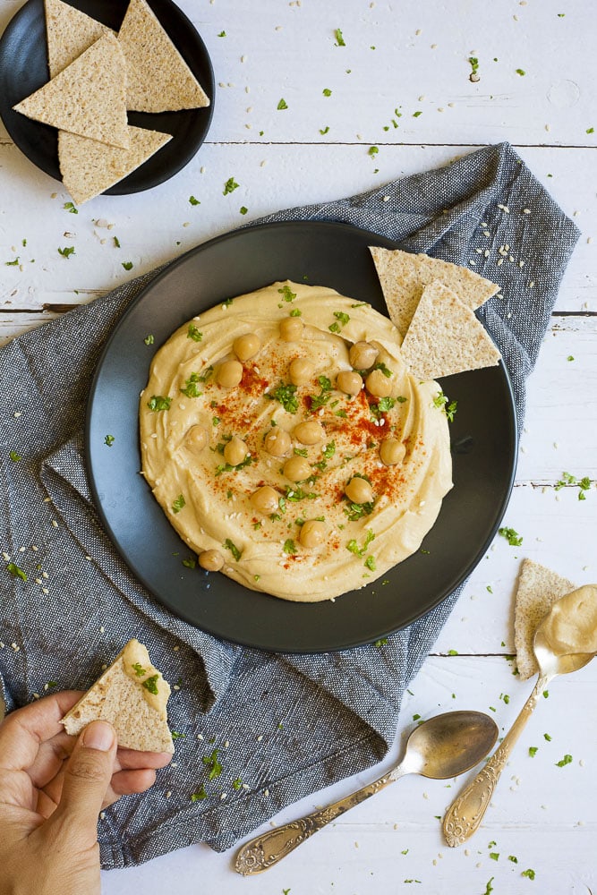 Black plate on grey tablecloth with oil-free hummus spread in the middle sprinkled with cooked chickpeas, fresh parsley, sweet paprika and sesame seeds.