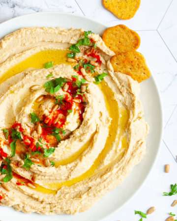 White plate with light brown hummus sprinkled on top with sunflower seeds, chopped parsley and paprika powder. Crackers are scattered around it.