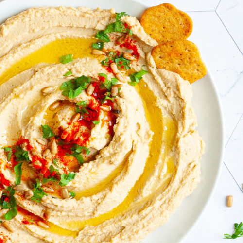 White plate with light brown hummus sprinkled on top with sunflower seeds, chopped parsley and paprika powder. Crackers are scattered around it.