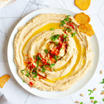 White plate with light brown hummus sprinkled on top with sunflower seeds, chopped parsley and paprika powder. Crackers are scattered around it.
