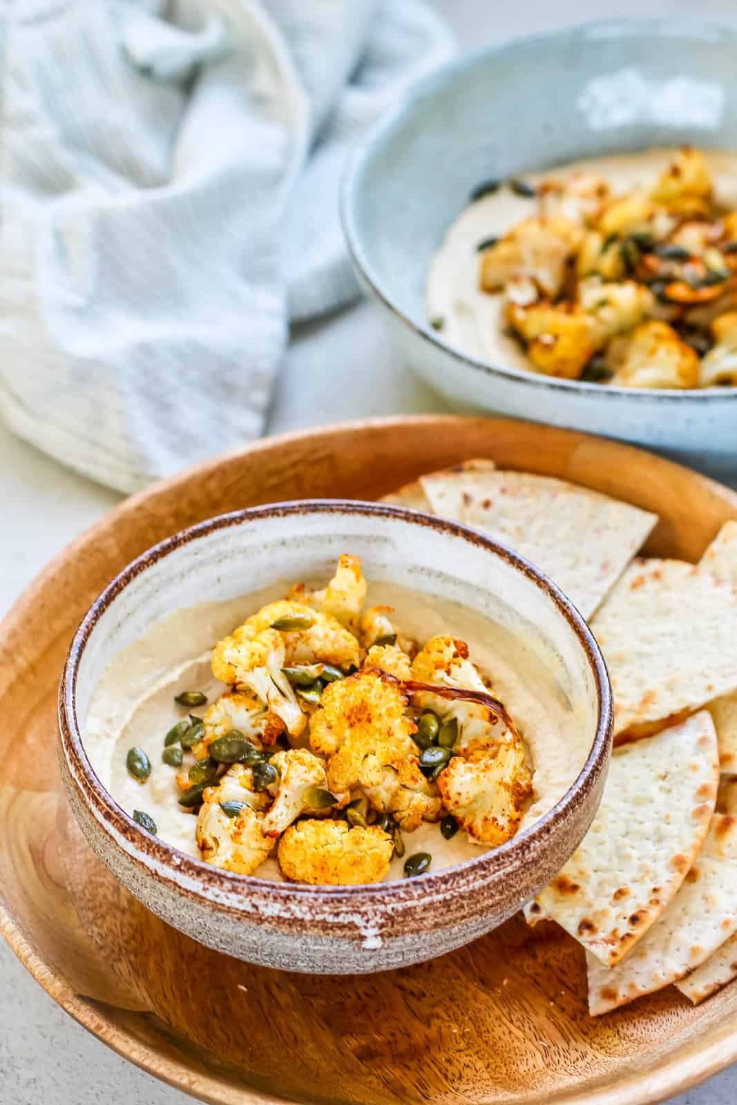 Light brown bowl with hummus topped with crispy yellow cauliflower florets and pumpkin seeds. Served with pita bread triangles on the side.