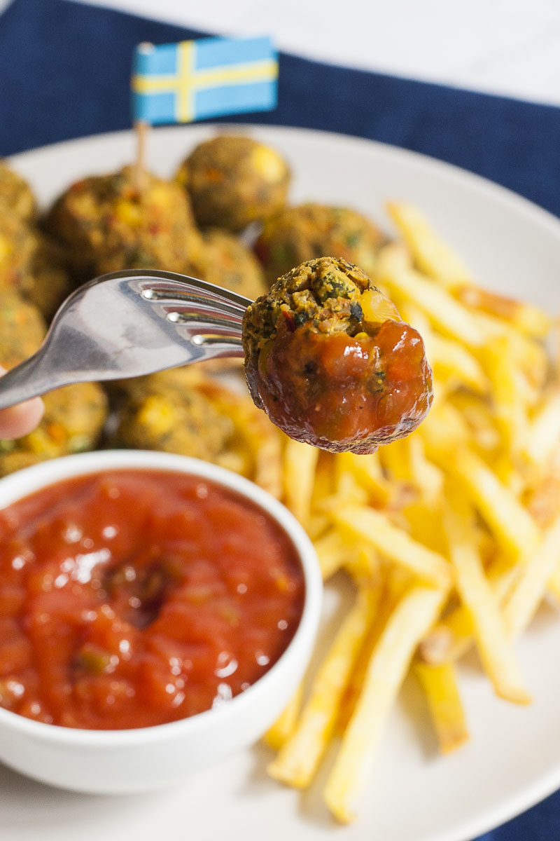 A fork is pierced through a brown veggie ball. In the background fried potato chips on a white plate and a small white bowl with red sauce. A blue yellow flag is in the background.