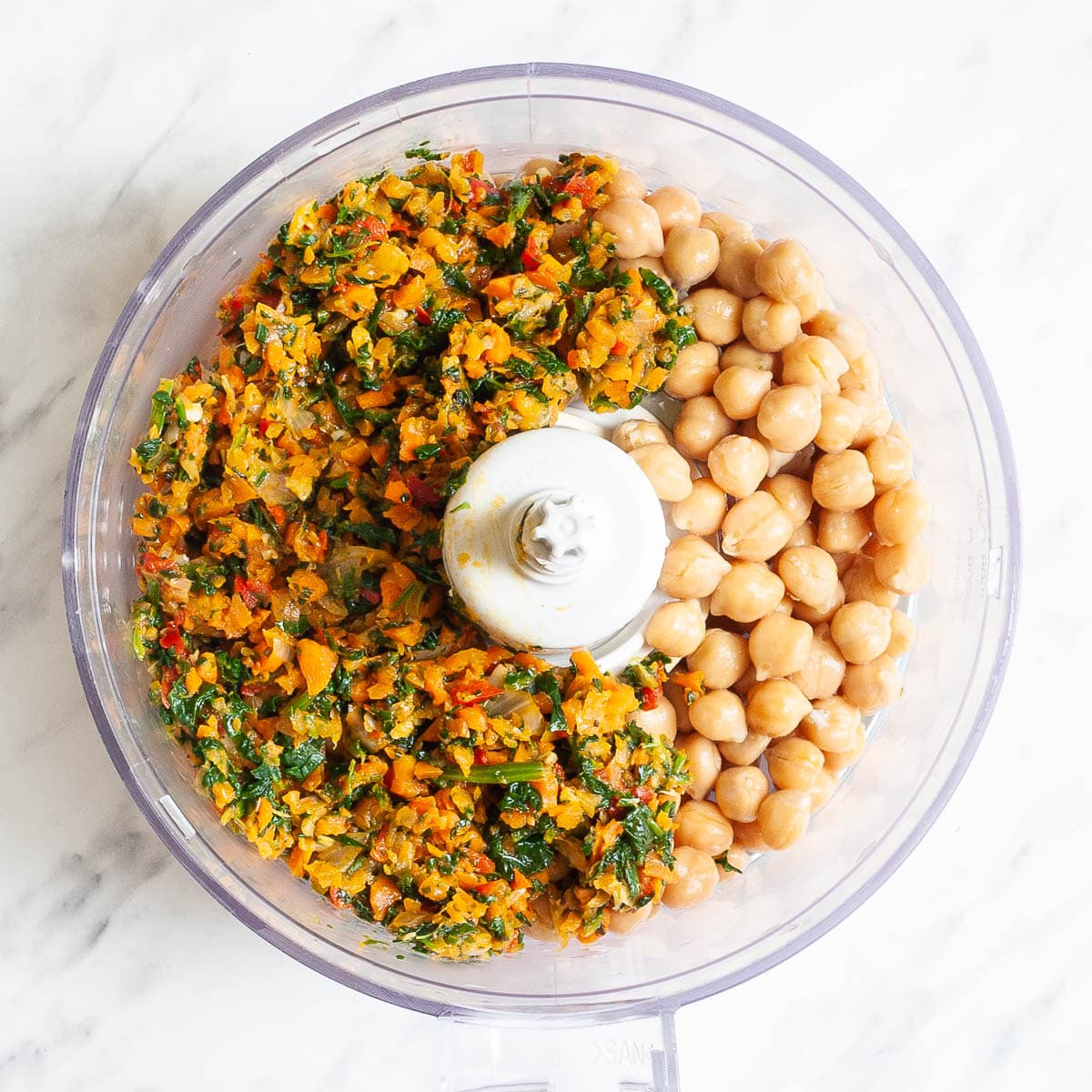 Food processor with chickpeas, finely chopped spinach, carrots, onion, red pepper, and garlic pieces.