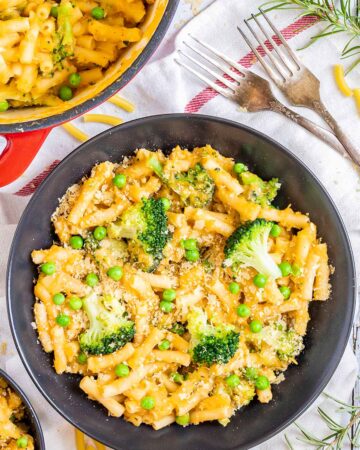 A red white enameled Dutch oven and 2 black bowls are serving short pasta in thick yellow sauce with broccoli florets and green peas added as toppings.