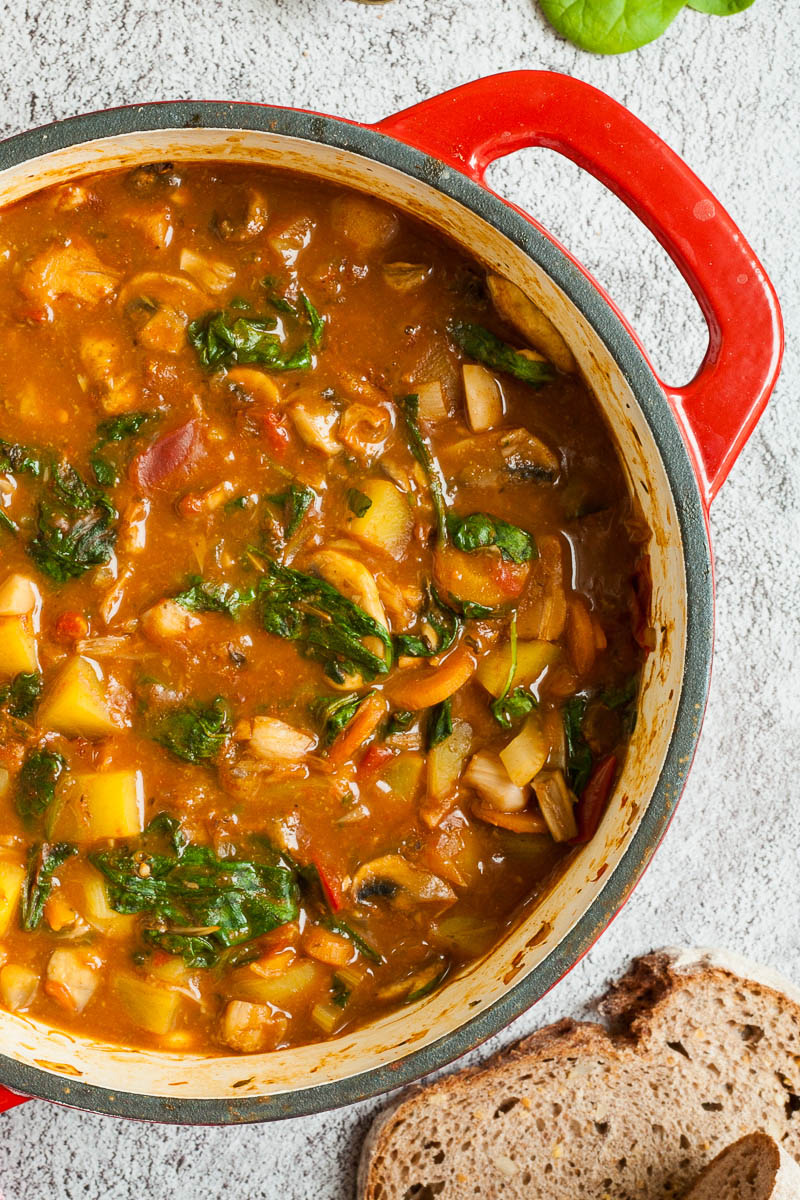 Red Dutch oven from above with a stew where you can see chopped potatoes, spinach leaves, carrot slices, tomatoes, and mushroom slices. Slices of bread is next to it.