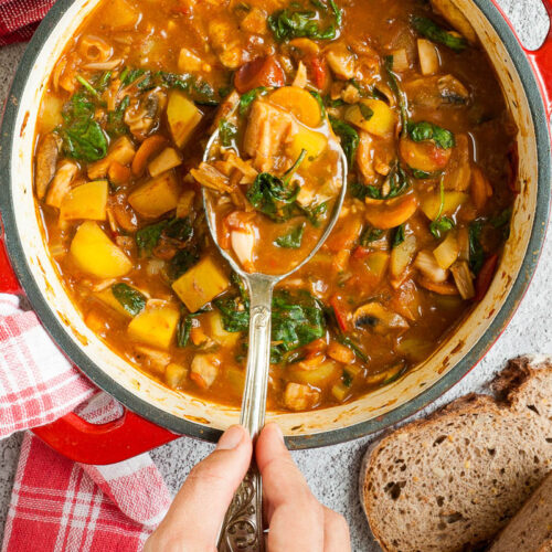 Red Dutch oven from above with a stew where you can see chopped potatoes, spinach leaves, carrot slices, tomatoes, and mushroom slices. Slices of bread is next to it. A hand is holding a spoon taking some from the middle
