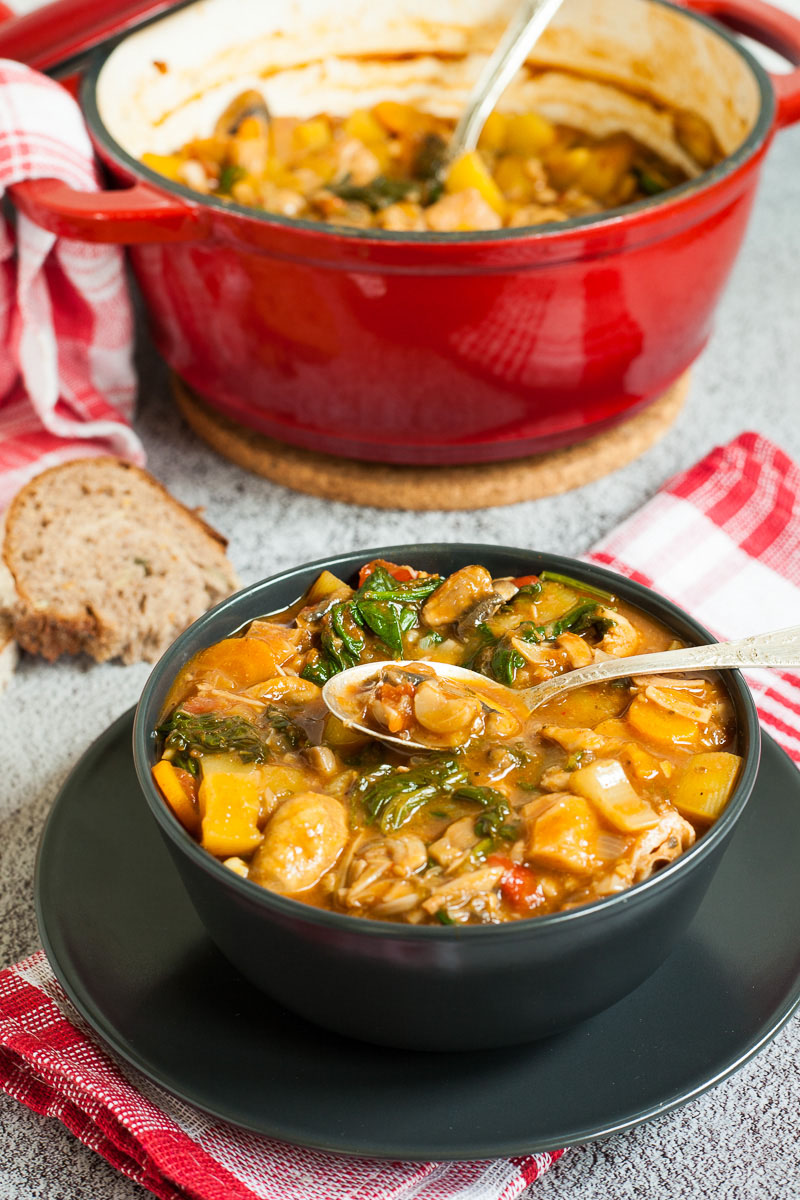 A black bowl with a stew where you can see chopped potatoes, spinach leaves, carrot slices, tomatoes, and mushroom slices. Slices of bread is next to it. A red Dutch oven is in the background with the remaining stew