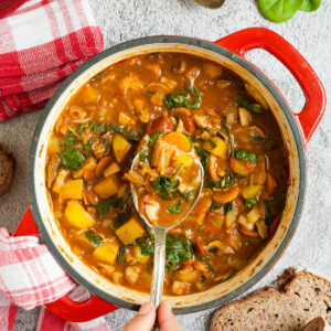 Red Dutch oven from above with a stew where you can see chopped potatoes, spinach leaves, carrot slices, tomatoes, and mushroom slices. Slices of bread is next to it. A hand is holding a spoon taking some from the middle
