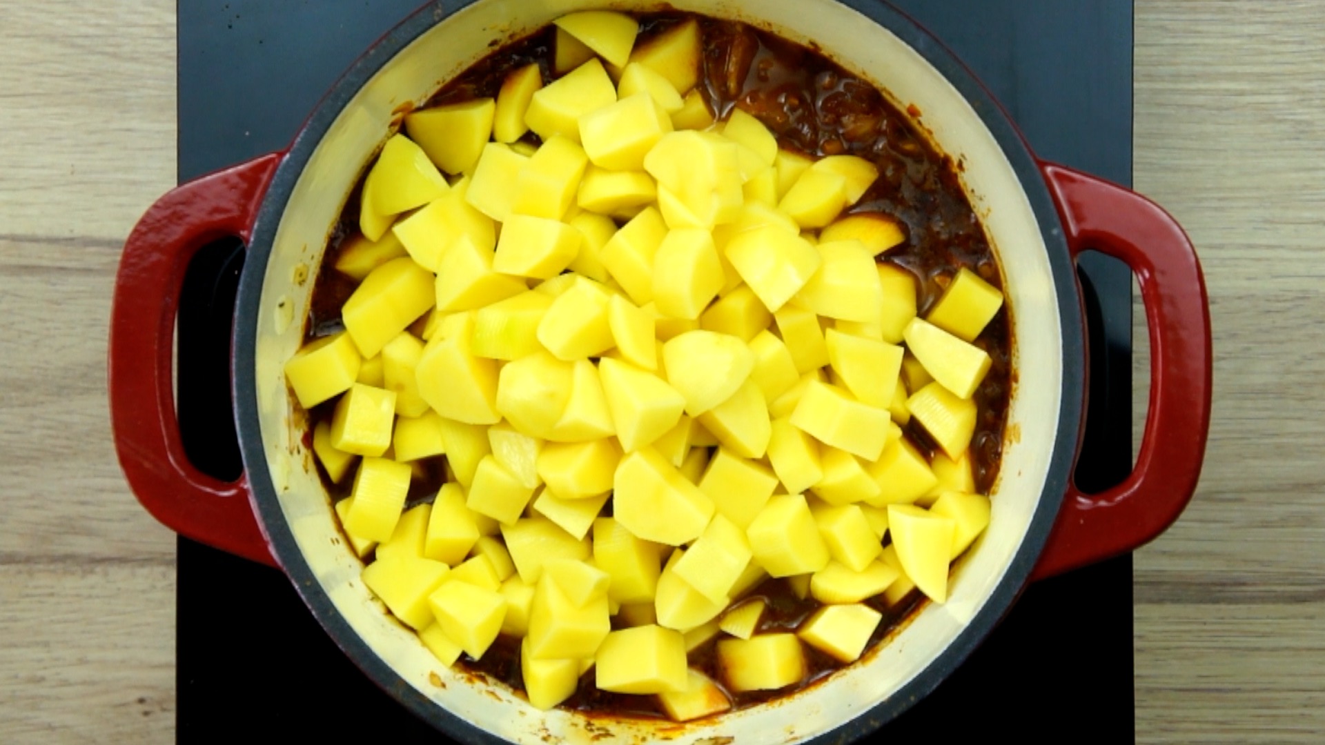 Red white Dutch oven with chopped onion, garlic, sliced carrots and jackfruit chunks in a brown broth covered with potato cubes.