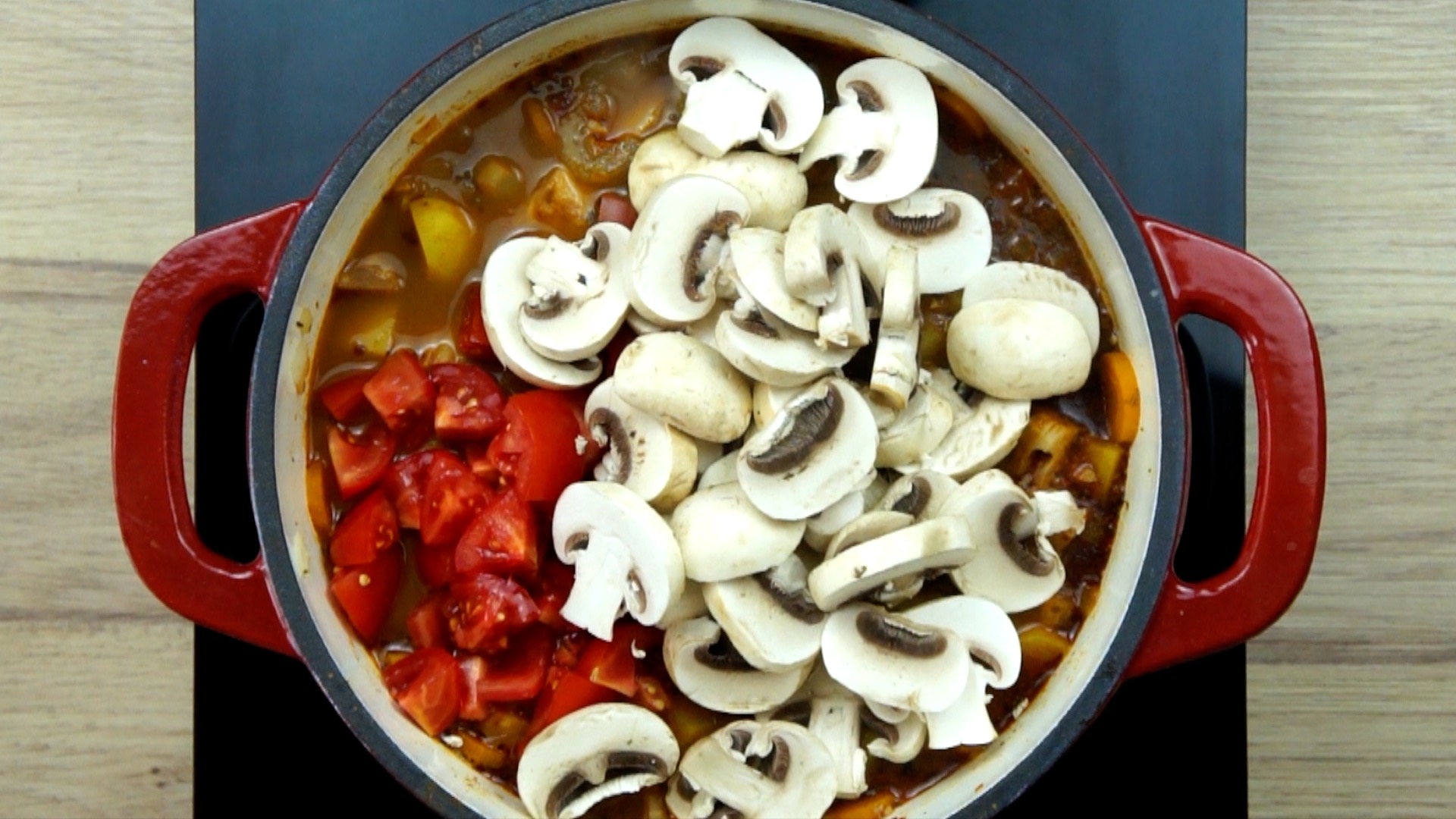 Red white Dutch oven with chopped onion, garlic, sliced carrots and jackfruit chunks in a brown broth covered with mushroom slices and tomato slices.