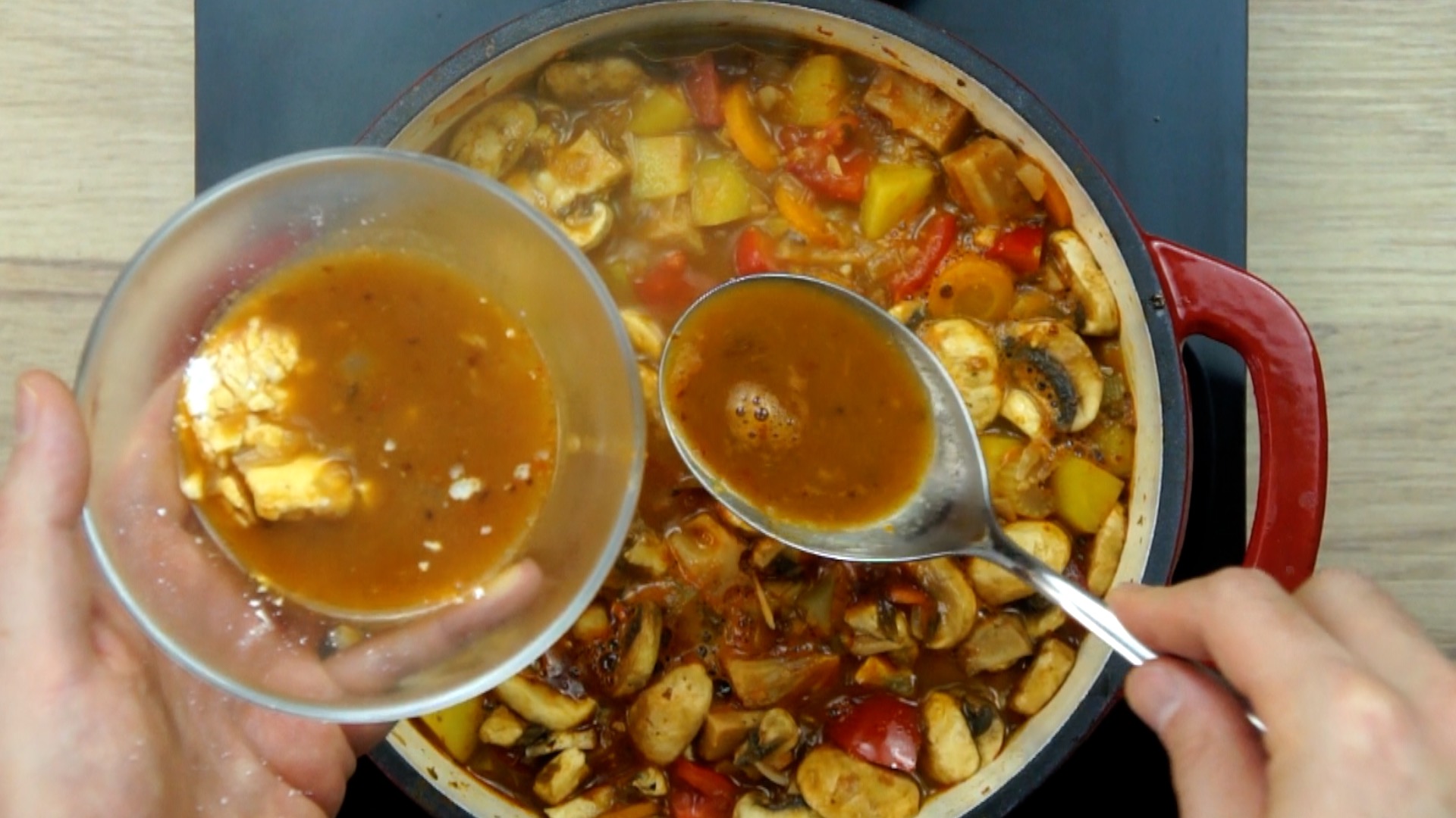 Red white Dutch oven with chopped onion, garlic, sliced carrots and jackfruit chunks in a brown broth. A hand is holding a glass bowl with white powder and adding a spoonful from the broth to it.