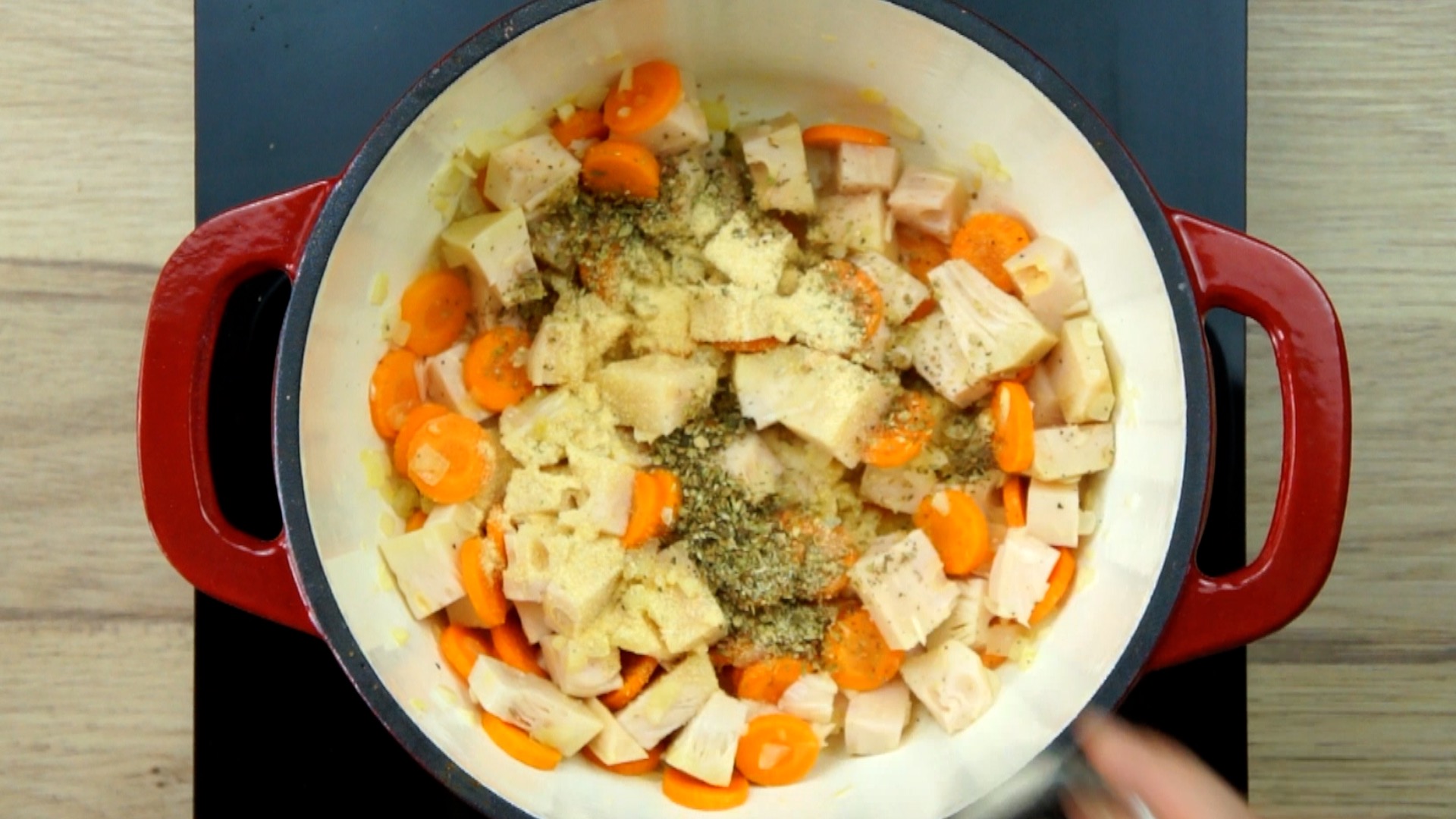 Red white Dutch oven with chopped onion, garlic, sliced carrots and jackfruit chunks topped with green dried herbs.
