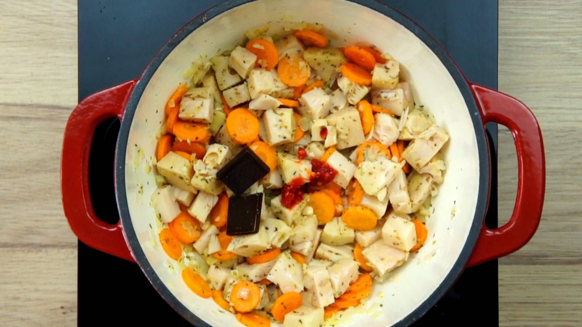 Red white Dutch oven with chopped onion, garlic, sliced carrots and jackfruit chunks topped with two chocolate cubes and a bit of red pepper.