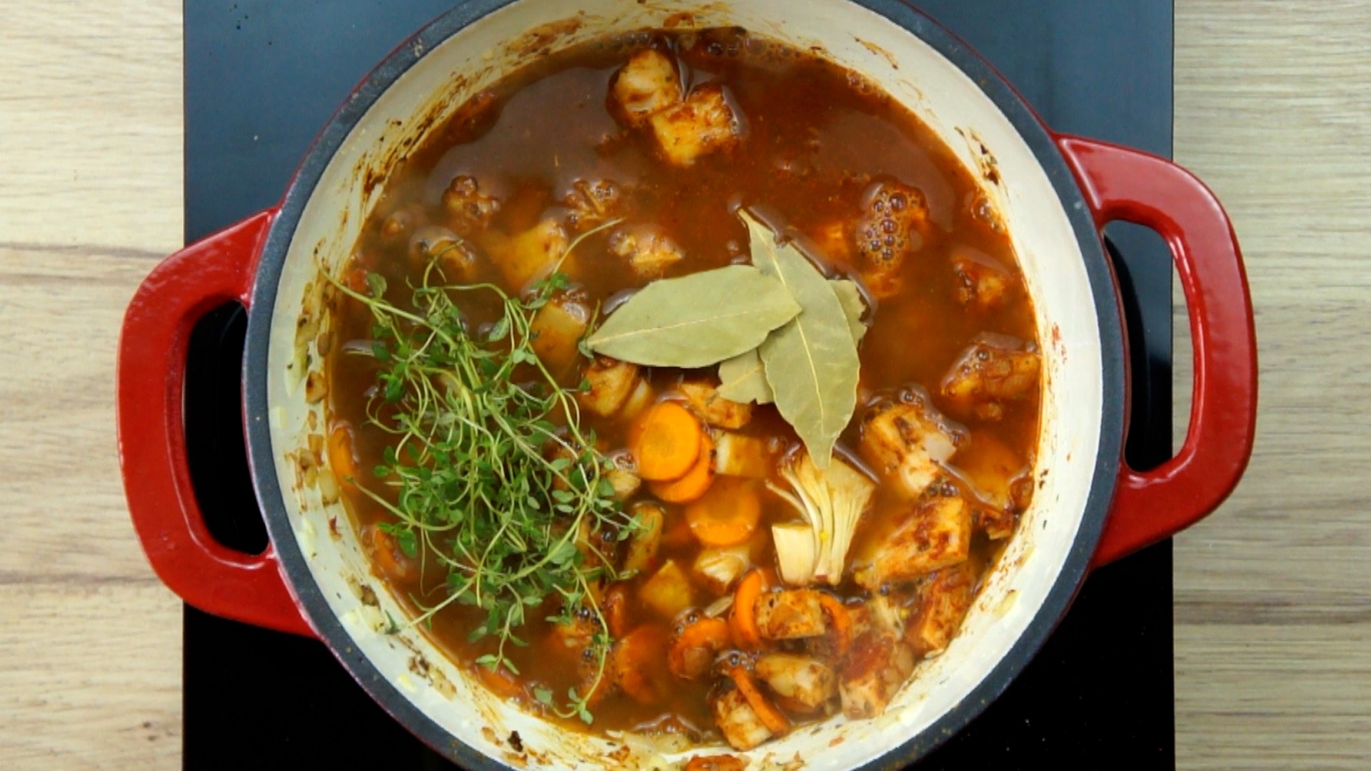 Red white Dutch oven with chopped onion, garlic, sliced carrots and jackfruit chunks in a brown broth with fresh herbs on top.