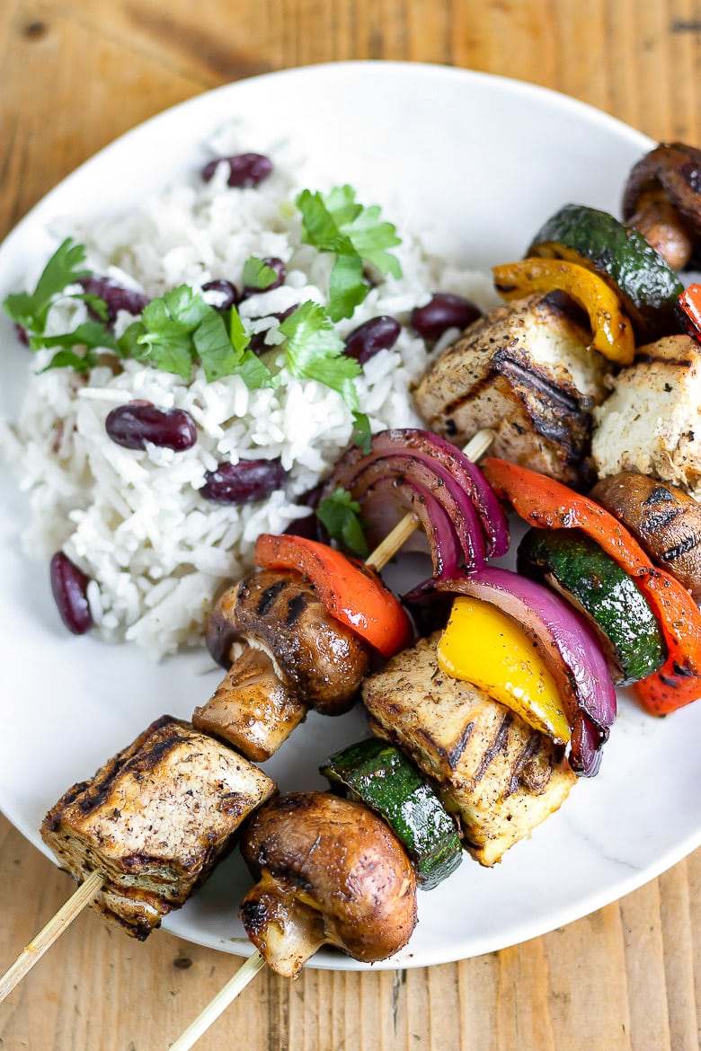 Two skewers with tofu cubes, mushroom, purple onion, zucchini, and red pepper are placed on w white plate with rice on the side.