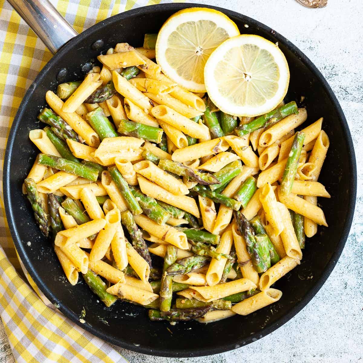 A frying pan with penne pasta, asparagus pieces and two lemon slices