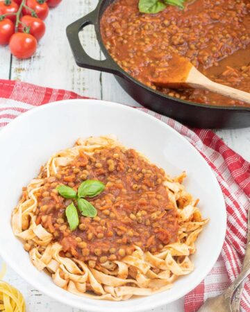 A white bowl of tagliatelle pasta topped with lentils in bolognese sauce. Leftover bolognese in the cast iron skillet is right next to it.