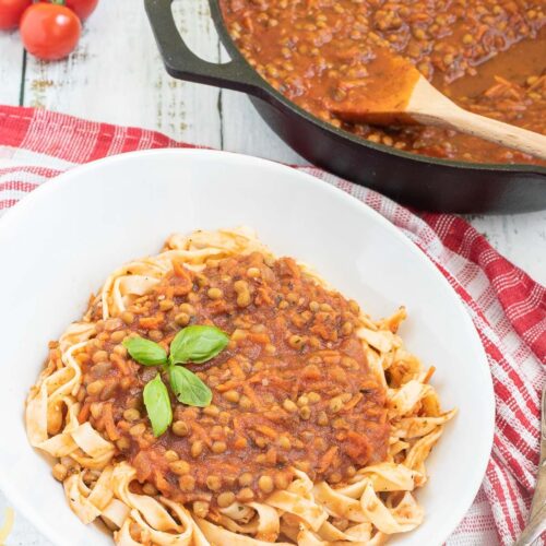 A white bowl of tagliatelle pasta topped with lentils in bolognese sauce. Leftover bolognese in the cast iron skillet is right next to it.