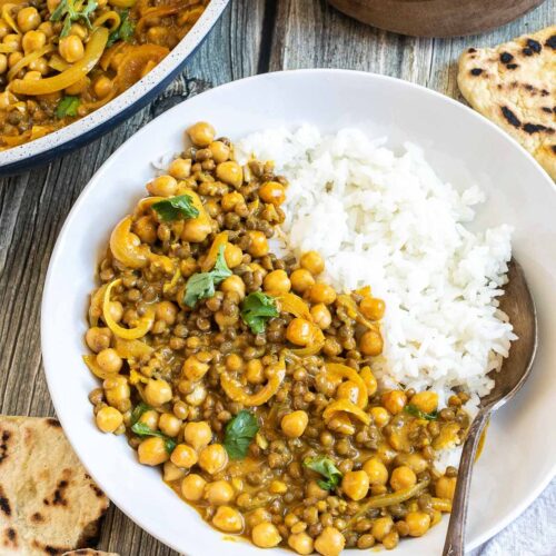 A white plate with white rice, chickpeas, and lentils, in a creamy light brown sauce sprinkled freshly chopped cilantro. A spoon is placed inside. Naan bread, a bowl of rice, and the remaining curry in the pan are next to it.
