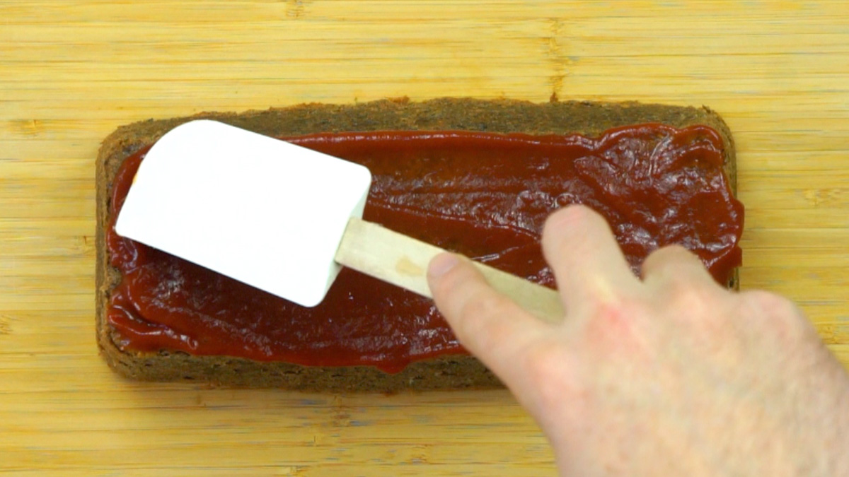 A hand is holding a spatula and spreading a red sauce on top of a brown lentil loaf.