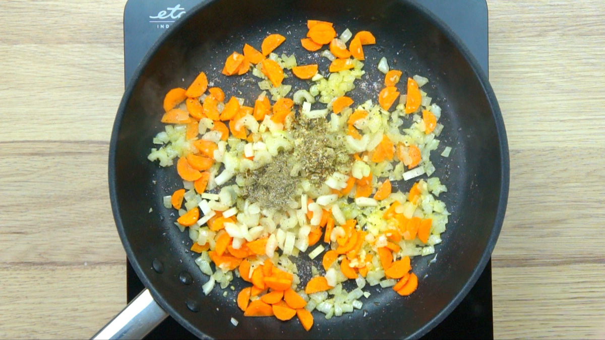 Black frying pan with minced onion, garlic, carrot, and celery pieces and a small heap of spices.