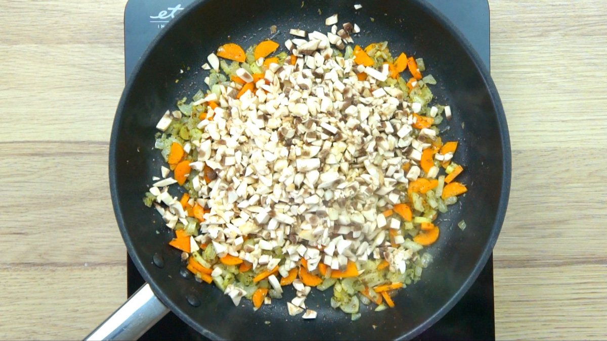 Black frying pan with minced onion, garlic, carrot, and celery pieces and a large heap of mushroom pieces.
