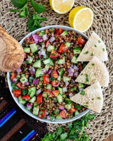 Large white bowl with a chopped mixed salad of brown lentils, cucumber, tomatoes, and red onion. 3 pita triangles are placed on the side.