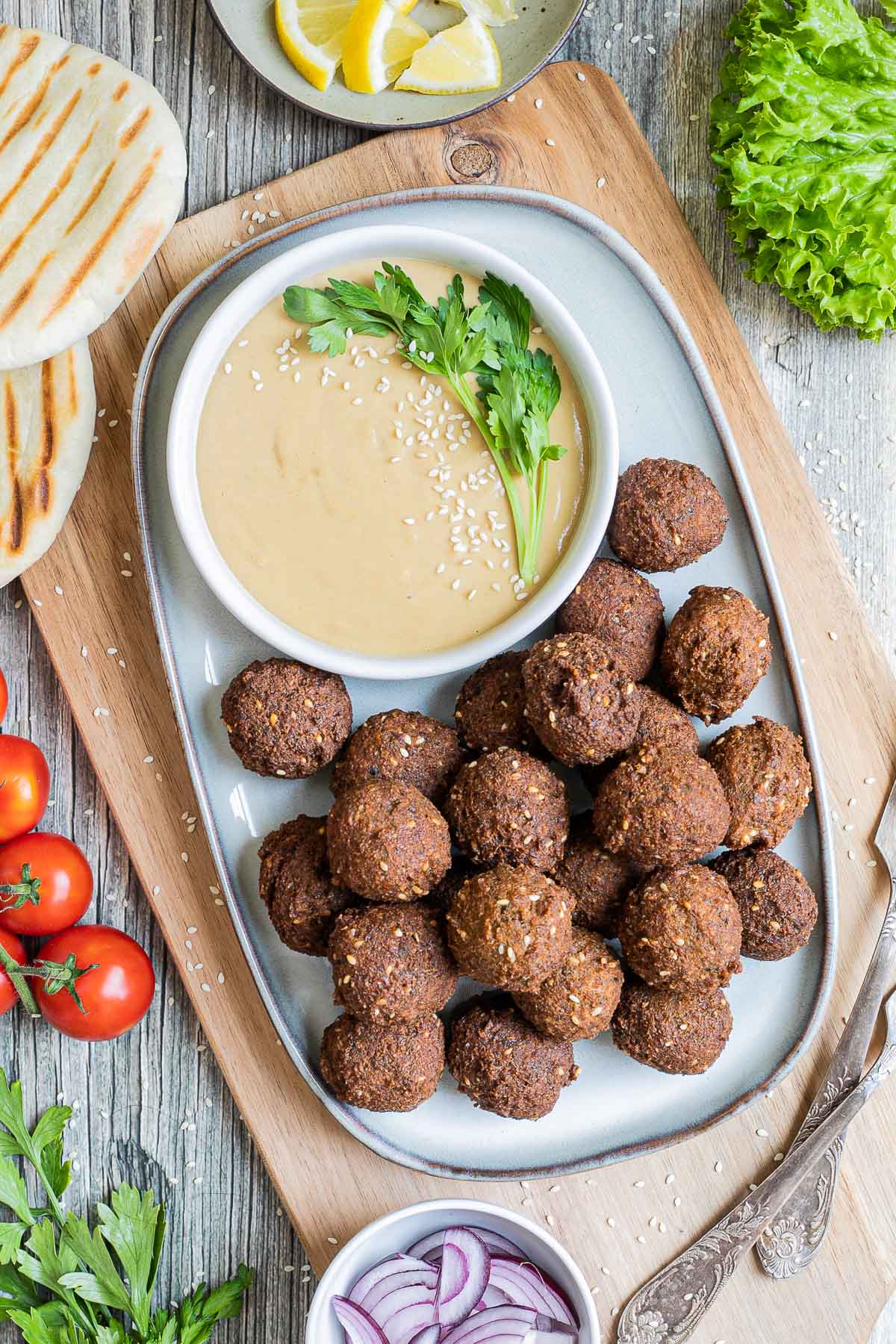 Light brown sauce in a white shallow bowl sprinkled with white sesame seeds and topped with a twig of parsley. Falafel balls, pite breads, and other raw vegetables are next to it.