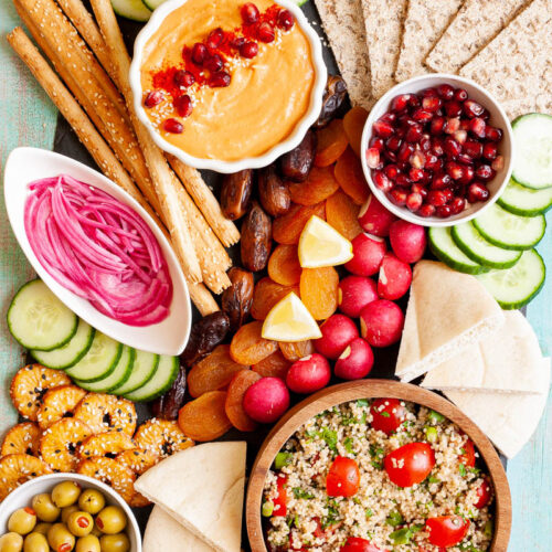 Black board with plenty of bowls, fruits and vegetables like cucumber, olives, dried apricot, pomegranate seeds, dates, orange hummus, tabbouleh salad