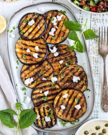 Large grilled slices of eggplant sprinkled with fresh herbs and crumbled cheese served on a light blue plate.