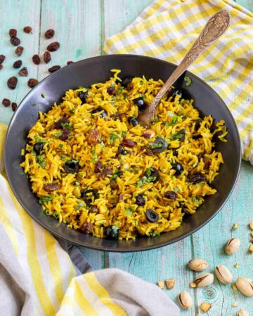 Yellow rice with raisins and pistachios and green herbs in a black bowl. More dry ingredients are scattered around it.