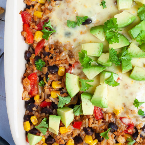 A ligth brown ceramic dish with colorful ingredients like black beans, corn, rice, melted cheese, bell pepper strips, chopped avocado, chopped green herbs.